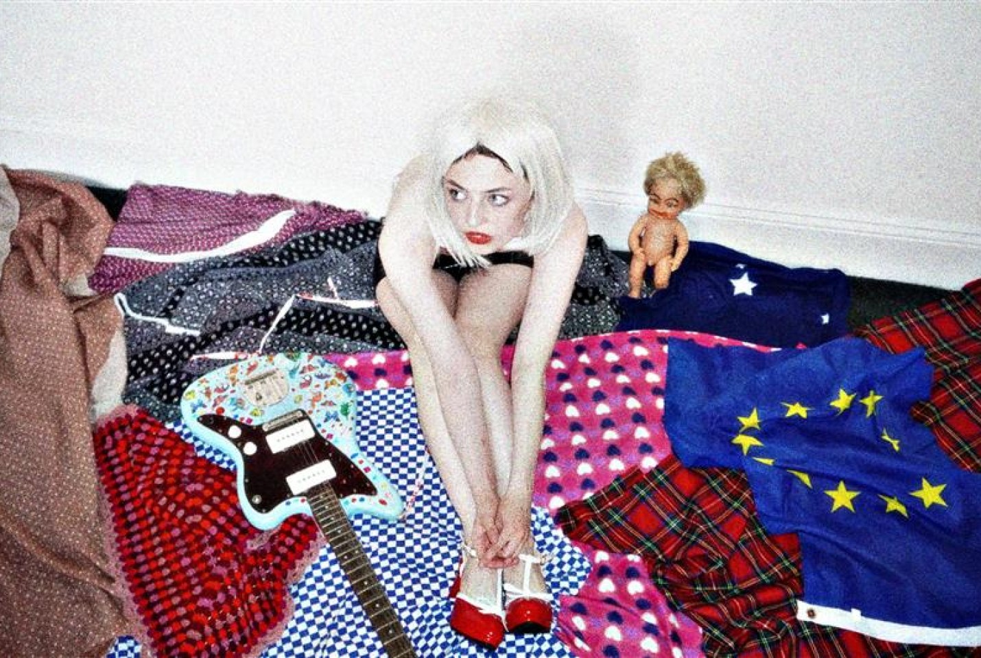 Girl in centre of image with blonde hair sitting on top of different blankets with a bass guitar to her left and a toy baby doll to her right.